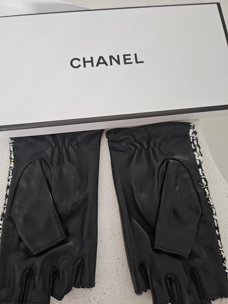 May include: Black leather fingerless gloves with white and black trim, and a white box with the word "CHANEL" printed in black. The gloves have a classic design.