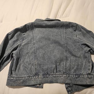 May include: A blue acid-wash denim jacket featuring a classic collar, long sleeves, and vertical seam detailing on the back. The garment has a cropped hemline with a structured waistband and visible stitching throughout the fabric construction.