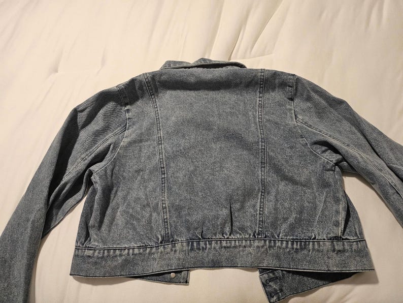 May include: A blue acid-wash denim jacket featuring a classic collar, long sleeves, and vertical seam detailing on the back. The garment has a cropped hemline with a structured waistband and visible stitching throughout the fabric construction.