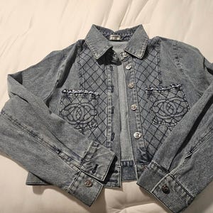 May include: A light blue denim jacket featuring a quilted diamond pattern on the front panels, two chest pockets with frayed edges and embroidered circular logos, a classic collar, and silver-toned metal buttons down the front and on the cuffs.
