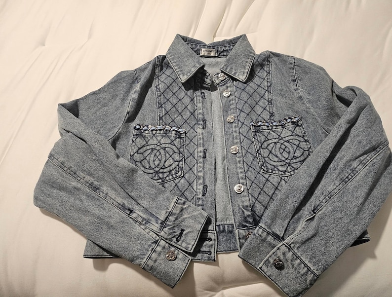 May include: A light blue denim jacket featuring a quilted diamond pattern on the front panels, two chest pockets with frayed edges and embroidered circular logos, a classic collar, and silver-toned metal buttons down the front and on the cuffs.