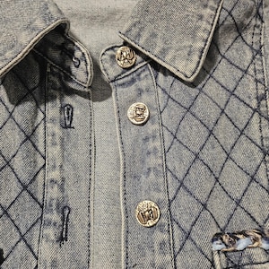 May include: A light blue denim jacket featuring a quilted diamond pattern across the fabric. The jacket has a classic collar, a button-down front with silver-toned metallic buttons, and a Chanel brand label stitched inside the neckline.