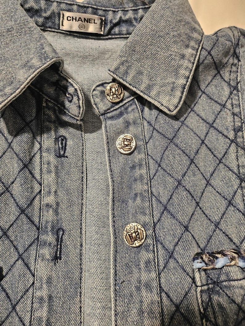 May include: A light blue denim jacket featuring a quilted diamond pattern across the fabric. The jacket has a classic collar, a button-down front with silver-toned metallic buttons, and a Chanel brand label stitched inside the neckline.