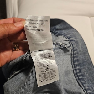 May include: A white fabric care label attached to a blue denim garment. The label displays material composition as 95% cotton and 5% polyester, along with multi-language washing and ironing instructions including 'wash on reverse' and 'do not iron on print'.