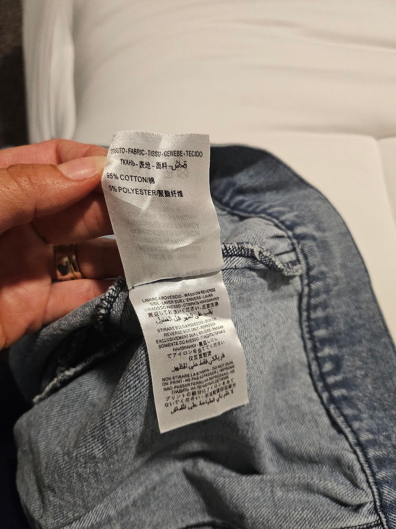 May include: A white fabric care label attached to a blue denim garment. The label displays material composition as 95% cotton and 5% polyester, along with multi-language washing and ironing instructions including 'wash on reverse' and 'do not iron on print'.