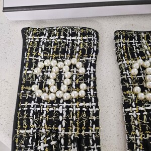 May include: A pair of black and white tweed gloves with gold accents. The gloves feature a pearl embellishment in the shape of a logo. The gloves have a fingerless design and a black trim.