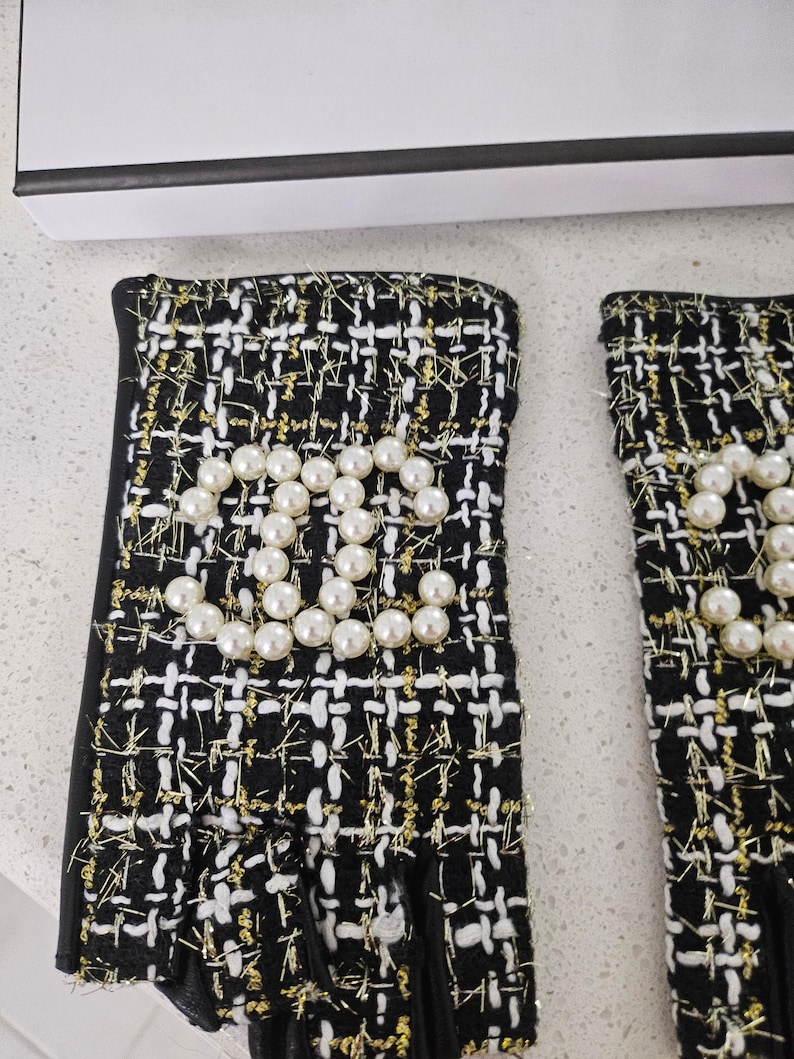 May include: A pair of black and white tweed gloves with gold accents. The gloves feature a pearl embellishment in the shape of a logo. The gloves have a fingerless design and a black trim.