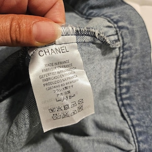May include: A white fabric garment tag attached to a blue denim textile. The tag features the brand name CHANEL in black text, followed by the phrase MADE IN FRANCE in multiple languages, and a series of five black laundry care symbols at the bottom.