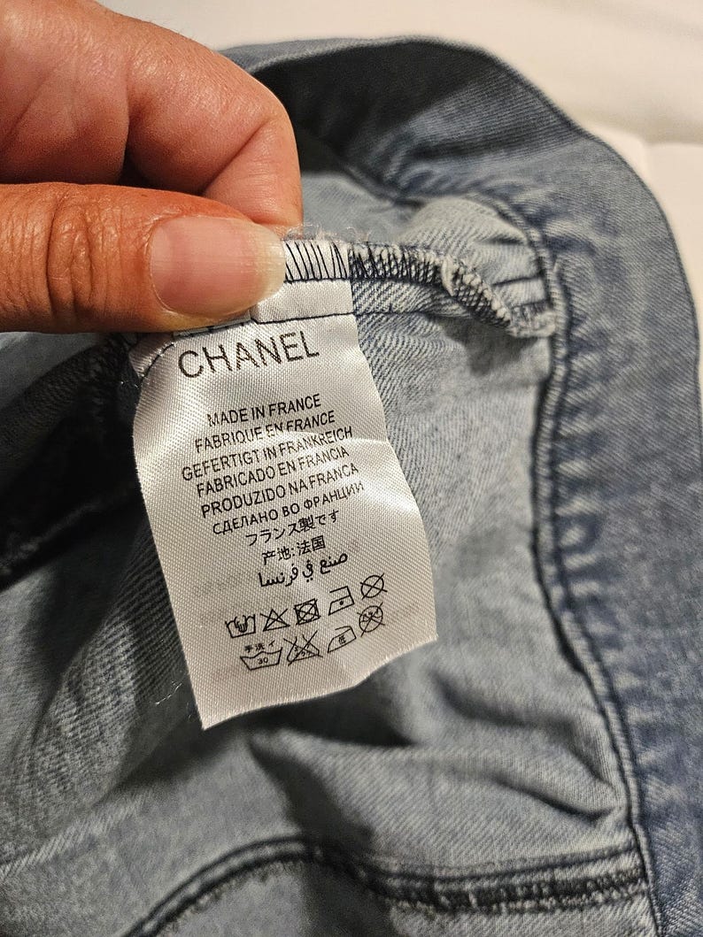 May include: A white fabric garment tag attached to a blue denim textile. The tag features the brand name CHANEL in black text, followed by the phrase MADE IN FRANCE in multiple languages, and a series of five black laundry care symbols at the bottom.