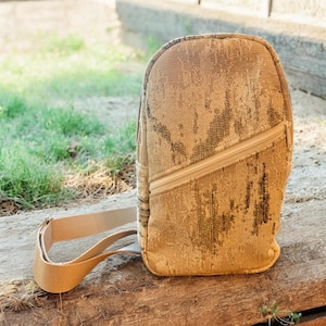 May include: A beige backpack with a diagonal zipper and a shoulder strap. The bag has a textured, patterned surface. The background includes green grass and a wooden surface.