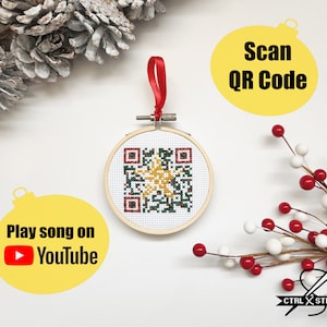 May include: A round, wooden ornament with a red ribbon and a green and gold cross-stitch QR code. The ornament is on a white background with a yellow ornament that says "Play song on YouTube" and a yellow ornament that says "Scan QR Code".