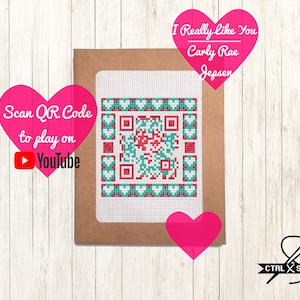 May include: A brown card with a white background featuring a cross-stitch design of a QR code surrounded by hearts. The text "Scan QR Code to play on YouTube" is in a pink heart shape on the left side of the card. The text "I Really Like You Carly Rae Jepsen" is in a pink heart shape on the right side of the card. The text "CTRL X STITCH" is in black text on a white background with a needle and thread design.
