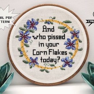 May include: A white embroidery hoop with a wooden frame. The embroidery features a floral wreath with purple flowers and green leaves surrounding the text "And who pissed in your Corn Flakes today?"