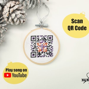 May include: A white cross-stitch ornament with a black and white QR code in the center. The ornament is framed in a wooden hoop and has a white ribbon attached. The ornament is surrounded by white pine cones and a yellow circle with the text "Scan QR Code".