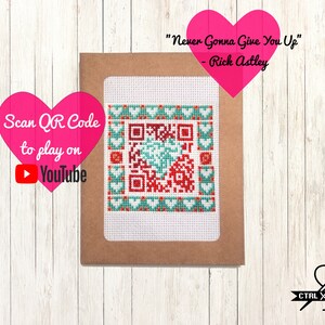 Op de afbeelding: Een bruine kaart met een wit kader rond een kruissteekontwerp van een QR-code met rode en groene harten. De tekst "Never Gonna Give You Up" - Rick Astley staat op de kaart gedrukt. De tekst "Scan QR Code to play on YouTube" staat op een roze hart gedrukt.