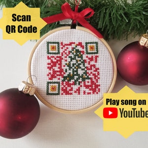 May include: A white Christmas ornament with a red and green cross-stitch design of a Christmas tree. The ornament has a QR code that can be scanned to play a song on YouTube. The ornament is surrounded by red Christmas ornaments.