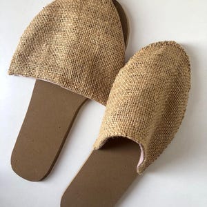 May include: A pair of natural-toned burlap slippers. The slippers have a brown sole and a woven burlap upper. The interior lining is a light pink color. The slippers are designed for indoor use and are placed on a white surface.