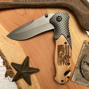 May include: A folding pocket knife with a black carbon fiber handle and a wood inlay featuring an American flag with a silhouette of a person holding a rifle. The blade is partially open and has a serrated edge.