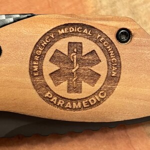 May include: A wooden knife handle with a laser engraved design. The design features a star of life symbol with the words "Emergency Medical Technician Paramedic" around it.