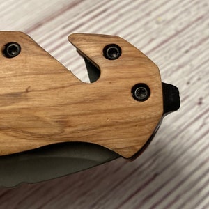 May include: Close-up of a wooden pocket knife handle with a black metal blade. The handle has a wood grain pattern and three black screws.