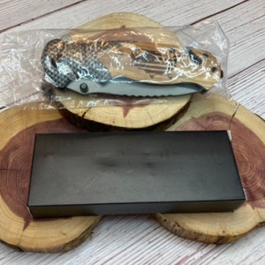 May include: A black folding knife with a wood handle and a carbon fiber pattern. The knife is in a plastic bag and is sitting on a wooden surface.