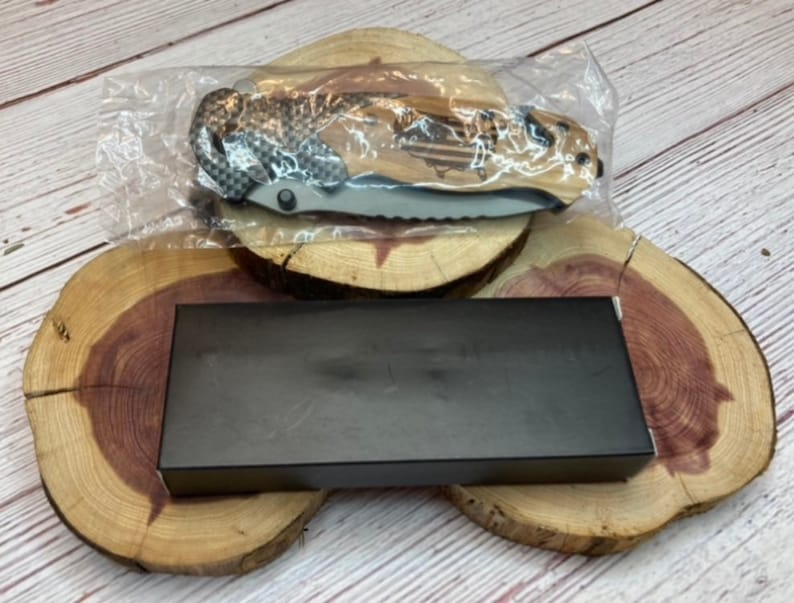 May include: A black folding knife with a wood handle and a carbon fiber pattern. The knife is in a plastic bag and is sitting on a wooden surface.