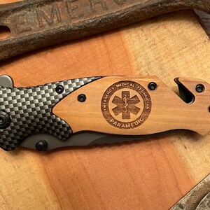 May include: A black and gray pocket knife with a wood handle. The handle has a laser engraved design that says "Emergency Medical Technician Paramedic" with a star of life symbol.
