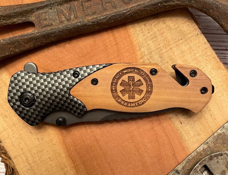 May include: A black and gray pocket knife with a wood handle. The handle has a laser engraved design that says "Emergency Medical Technician Paramedic" with a star of life symbol.