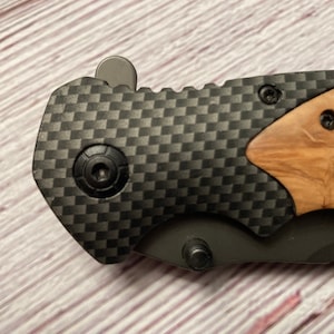 May include: A black folding knife with a carbon fiber handle and a wood inlay. The knife has a black blade and a silver button on the handle.