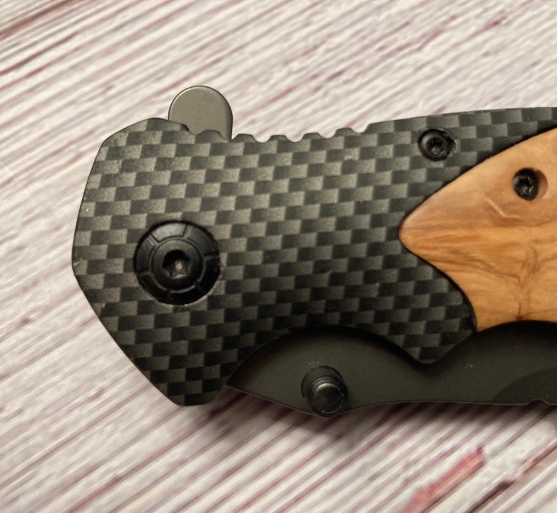May include: A black folding knife with a carbon fiber handle and a wood inlay. The knife has a black blade and a silver button on the handle.