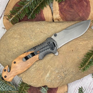 May include: A black and brown pocket knife with a wood handle and a safety feature. The knife is on a brown rock with green foliage.