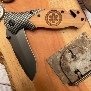 May include: A black and wood folding pocket knife with a serrated blade and a wood handle. The handle has a laser engraved design that says "Emergency Medical Technician Paramedic" with a medical symbol.