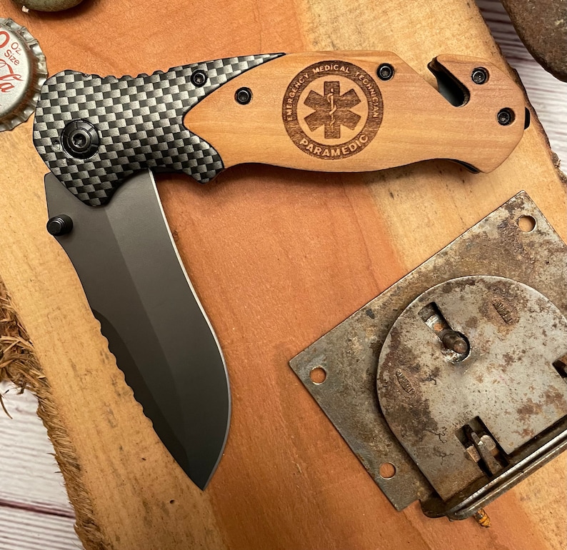 May include: A black and wood folding pocket knife with a serrated blade and a wood handle. The handle has a laser engraved design that says "Emergency Medical Technician Paramedic" with a medical symbol.