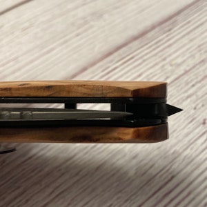 May include: Close-up of a wooden handled pocket knife with a black blade. The blade is partially open and the tip is visible.