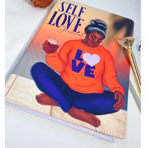 Pu&ograve; includere: Diario di Autoamore con un'illustrazione colorata di una donna seduta con un bicchiere di vino. La donna indossa una camicia arancione con le parole "Love" in lettere viola e un cuore bianco sul davanti. La copertina del diario ha uno sfondo bianco con un motivo a venature di legno marrone chiaro.