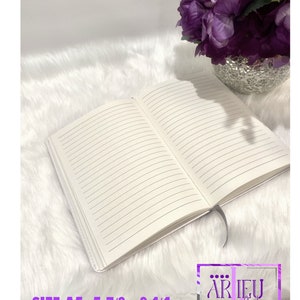 Pu&ograve; includere: Un quaderno bianco con pagine a righe, aperto per rivelare le pagine vuote. Il quaderno &egrave; su una superficie bianca e pelosa. Il testo "SIZE A5 5 7/8 - 8 1/4" &egrave; visibile nella parte inferiore dell'immagine. Un arrangiamento di fiori viola &egrave; visibile nell'angolo in alto a destra dell'immagine.