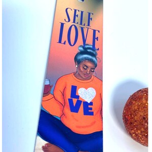 Pu&ograve; includere: Un segnalibro con una donna con un bicchiere di vino, che indossa una felpa arancione brillante con la parola "LOVE" in lettere blu e un cuore bianco. Lo sfondo &egrave; un tramonto con le parole "SELF LOVE" in lettere blu in alto.