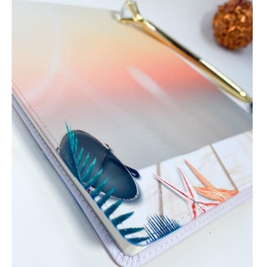 Pu&ograve; includere: Un taccuino bianco con un design ombr&eacute; colorato sulla copertina. La copertina presenta una borsa blu, foglie verdi e uccelli arancioni e rossi. Il taccuino ha un bordo cucito bianco.