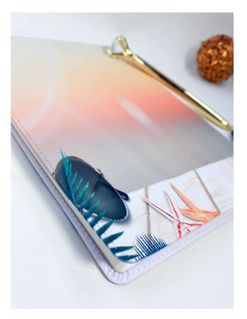 Pu&ograve; includere: Un taccuino bianco con un design ombr&eacute; colorato sulla copertina. La copertina presenta una borsa blu, foglie verdi e uccelli arancioni e rossi. Il taccuino ha un bordo cucito bianco.