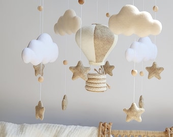 May include: A wooden baby mobile with a beige and white hot air balloon, beige and white clouds, and beige stars. The mobile is hanging from a wooden ring with the text "JOSH KRONK 201-6" on it.