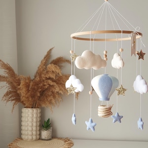 Op de afbeelding: Een hangende mobiel met een rond houten frame en daaraan bevestigde vilten elementen, waaronder een lichtblauwe en witte heteluchtballon, diverse witte wolken, gouden glittersterren, kleine houten kralen en lichtblauwe sterren.