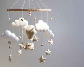 May include: A baby mobile featuring a hot air balloon, clouds, and stars. The mobile is suspended from a wooden frame with a string. The hot air balloon is beige and white, and the clouds and stars are white and beige. The text "OSIM SKRIVK 201-6" is printed on the wooden frame.