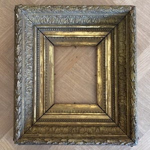 May include: A rectangular picture frame featuring an ornate, multi-layered design with a weathered gold and silver finish. The frame displays intricate relief carvings of leaf patterns and beaded detailing along its deep, stepped inner and outer borders.