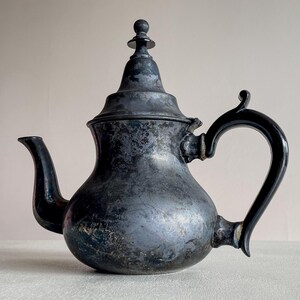 Peut inclure: Théière ancienne de couleur argentée avec un bec verseur incurvé et une grande anse arquée. Le couvercle décoratif est surmonté d'un petit bouton. La surface montre des signes d'âge et d'usure.