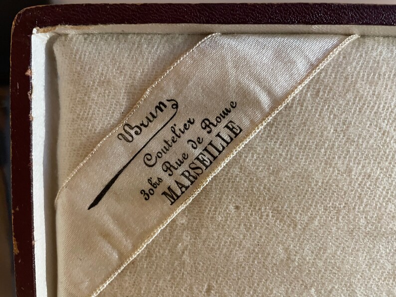 May include: A white satin ribbon with black text that reads "Brums Coutelier 30 bis Rue de Rome MARSEILLE".