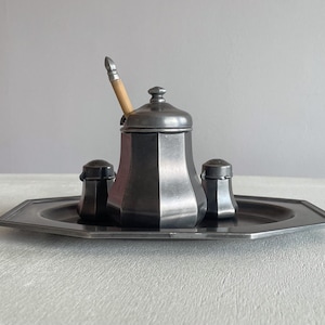 Puede incluir: Un juego de condimentos de peltre con un azucarero con cuchara de madera y dos saleros y pimenteros. El juego se encuentra sobre una bandeja octogonal. El metal tiene un acabado oscuro y mate.