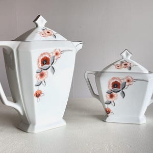 Peut inclure: Service à thé en porcelaine blanche composé d'une théière et d'un sucrier. Chaque pièce présente un motif floral avec des accents rouges, orange et gris. L'ensemble a une forme géométrique et une bordure argentée.