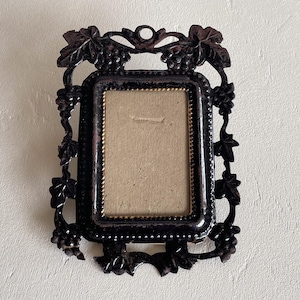 May include: Black rectangular picture frame with grape vine and leaf detailing. The frame has a small, rectangular opening with a light tan background. The frame is made of a dark, possibly metal material.
