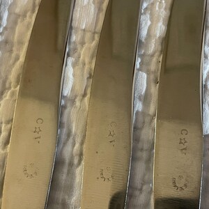 May include: Close-up of five silver-colored butter knives with a textured surface. Each knife has a star and the letters "C*1" engraved on the handle.