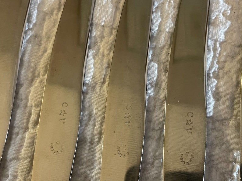 May include: Close-up of five silver-colored butter knives with a textured surface. Each knife has a star and the letters "C*1" engraved on the handle.